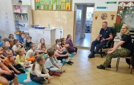 Grupa dzieci siedzących na dywanie słucha funkcjonariuszy Policji i Straży Granicznej na temat  bezpiecznych wakacji.