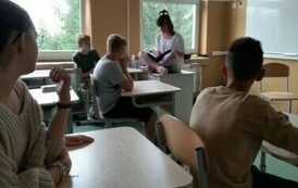 Uczniowie siedzą na krzesełkach, przy łstolikach szkolnych. Jedna osoba siedzi na biurku nauczyciela.