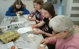 Uczennice i seniorka siedzą przy stole i malują jajka.
