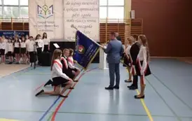 Uczniowie w galowych strojach z przewieszonymi biało-czerwonymi szarfami  klęczą przed sztandarem szkolnym trzymanym przez dyrektora szkoły.
