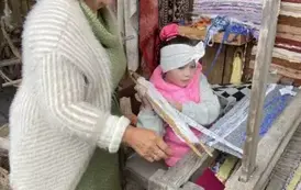 Pani stoi obok dziewczynki, kt&oacute;ra siedzi przy krosnach