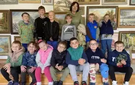 Dzieci stoją i siedzą wok&oacute;ł pani. Pozują do zdjęcia w galerii obraz&oacute;w