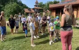 Dzieci z rodzicami stoją na trawie i słuchają co m&oacute;wi pani