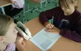 Uczennice siedzą w ławce i wykonują zadanie.