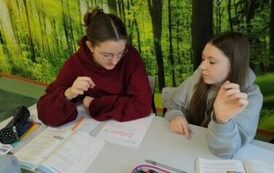 Uczennice siedzą w ławce i wykonują karty pracy z liczba 12.