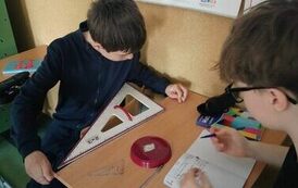 Uczniowie siedzą w ławce  i mierzą długość podstawki