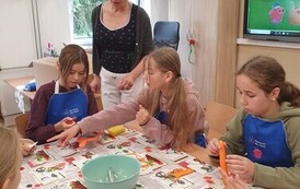 Dziewczynki siedzą przy stole i przygotowują sałatkę z marchewką, obok stoi kobieta w czarbej bluzce i białych spodniach.jpeg