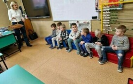 Siedmiu chłopc&oacute;w siedzi na piankowej kanapie, obok nich stoi nauczycielka z koszykiem